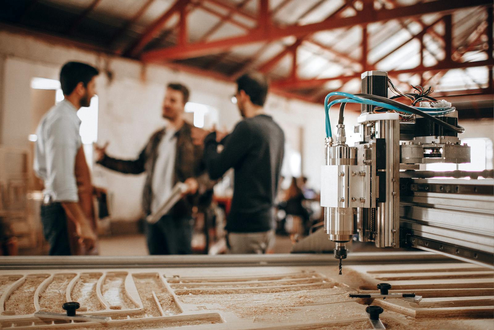 Three men discussing work in a woodworking shop with CNC machinery in focus.